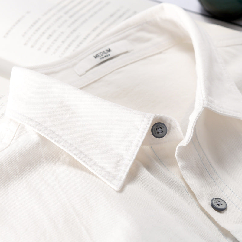 Camicia bianca in cotone con colletto classico e bottoni grigi, su sfondo di pagine aperte. Elegante abbigliamento maschile, moda sostenibile.
