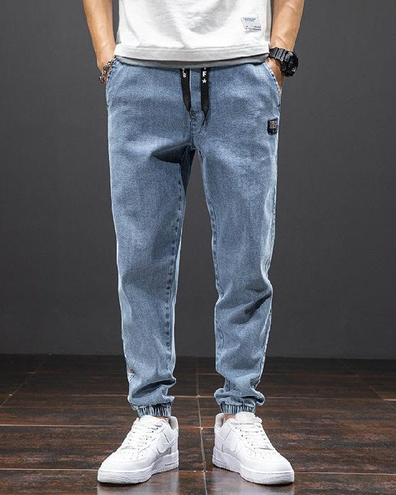 Jeans jogger uomo in denim blu chiaro, con coulisse nera, abbinati a scarpe da ginnastica bianche su pavimento in legno scuro.