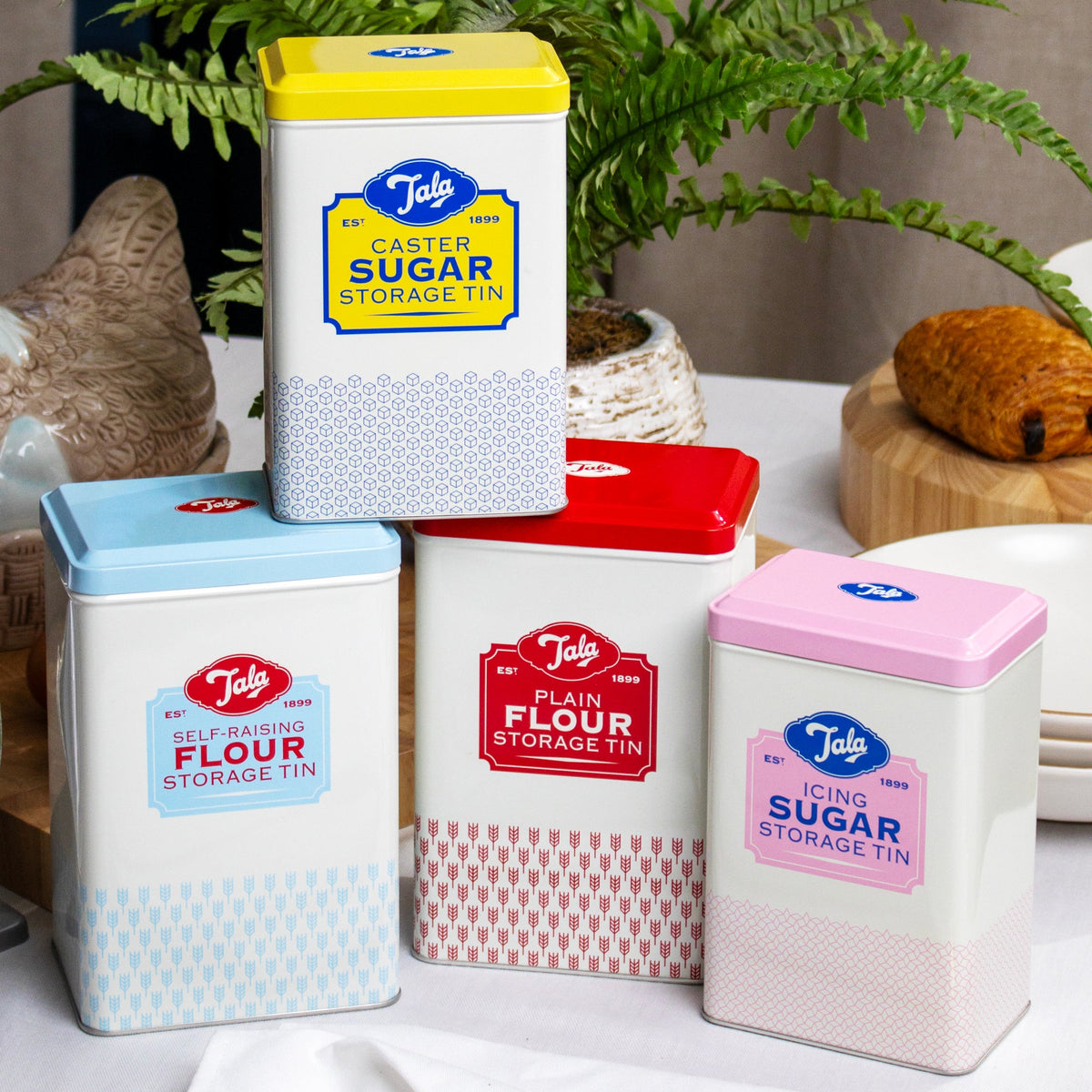 A set of four retro Tala branded storage tins with hinged lids, in various colors, labeled for flour and sugar storage, displayed on a kitchen counter.